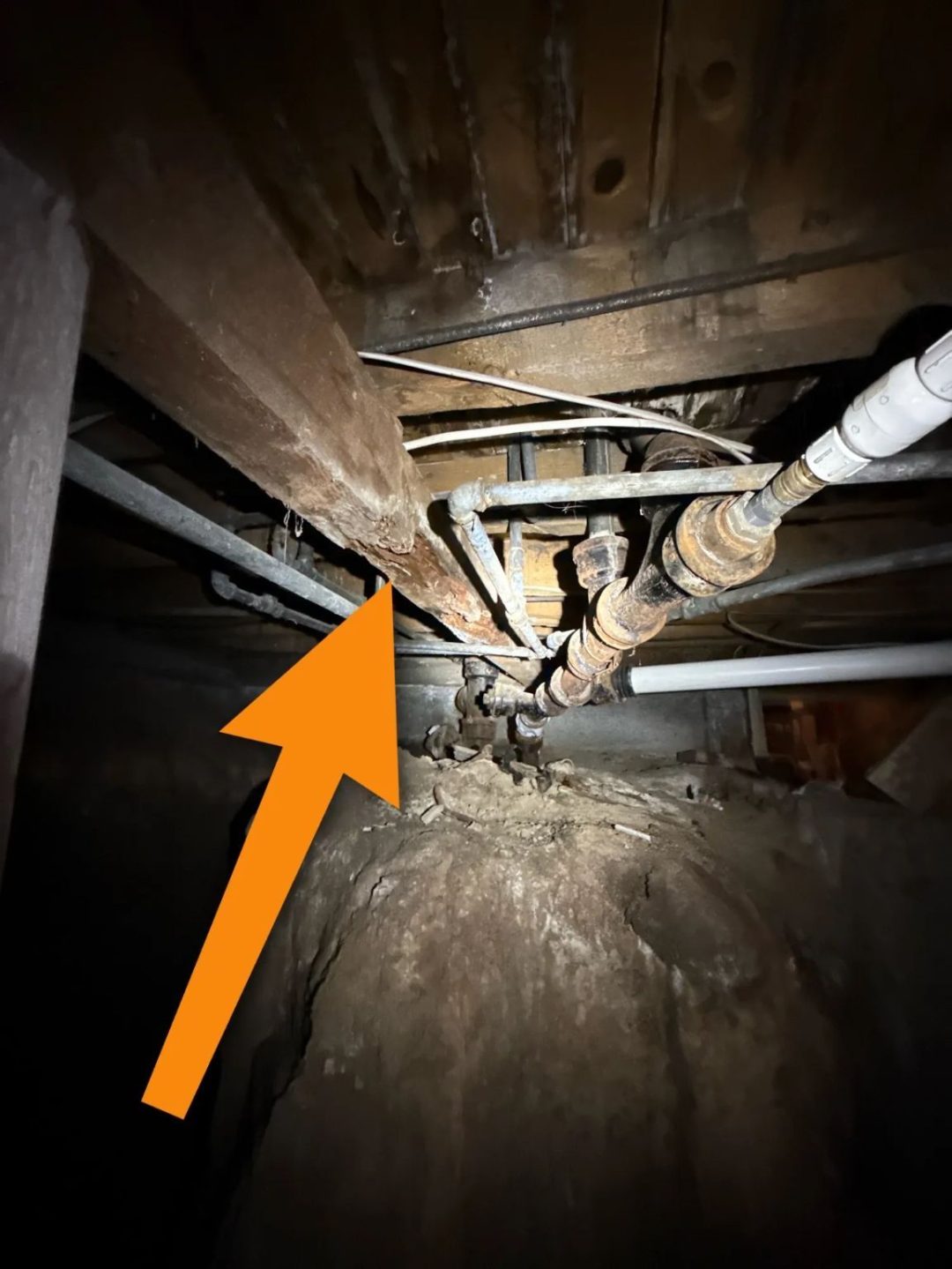 Crawl space under a Ronan, MT home showing corroded galvanized plumbing, deteriorated wood framing highlighted by an orange arrow, and a dirt floor with mixed pipe materials